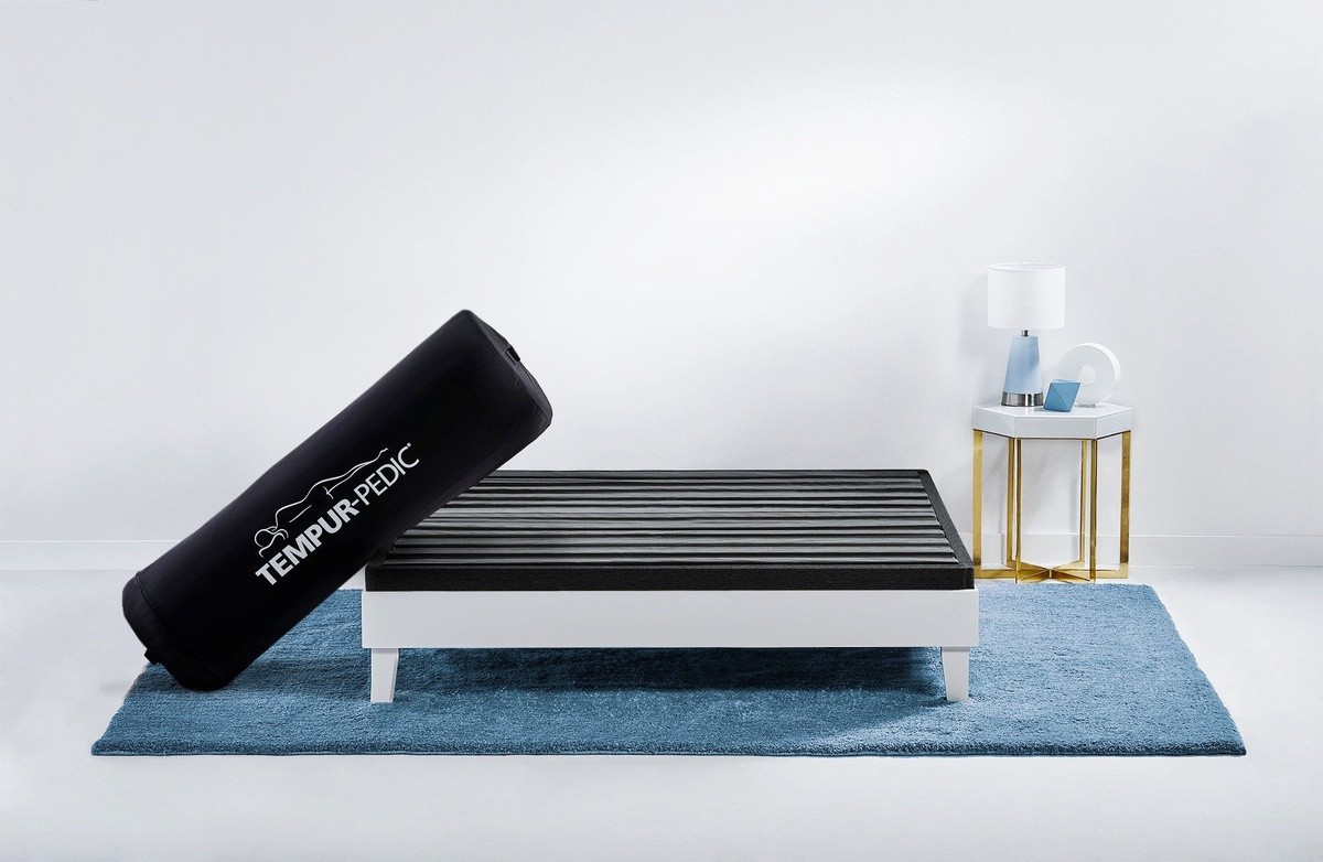 compressed and rolled up Tempur-Pedic Cloud mattress in a bag leaning against bare bedframe on a blue rug