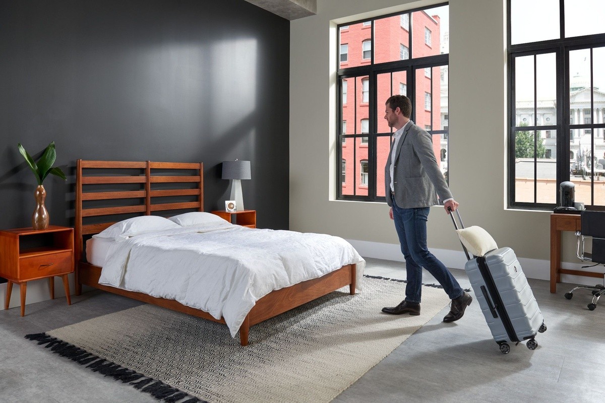 Man rolling a suitcase into a decorated bedroom with a wooden headboard and nightstands