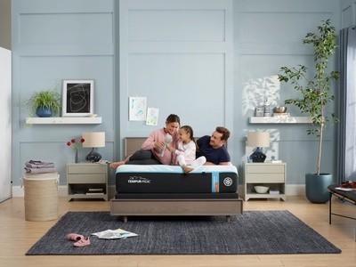 Happy family on a LuxeBreeze mattress sitting on a wooden frame in a decorated room with blue walls, a dark rug, and plant in the right corner