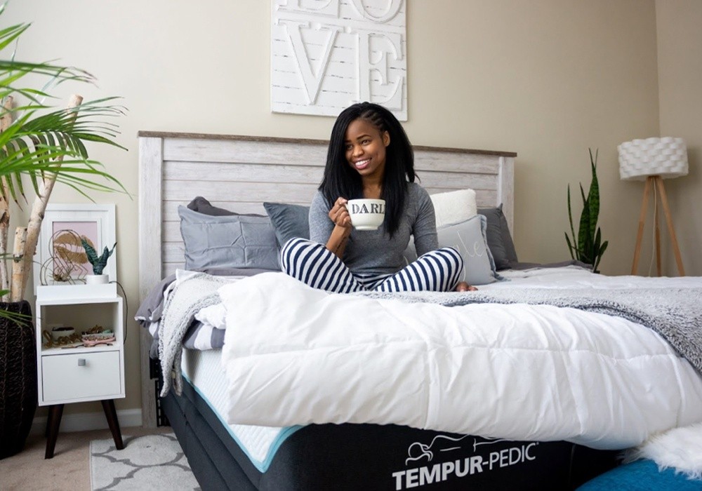 A woman sitting on a Tempur-Pedic mattress drinking out of a mug