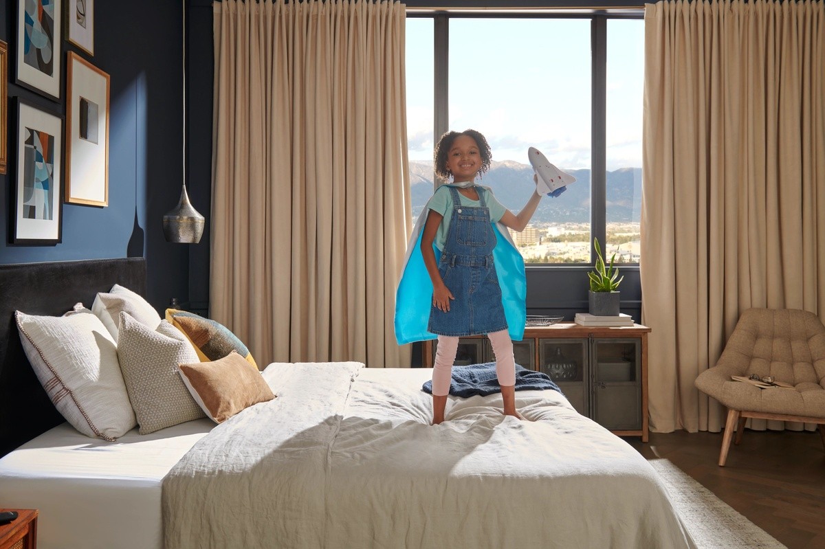 Child standing on a made bed in a decorated room wearing a cape and holding a toy rocket