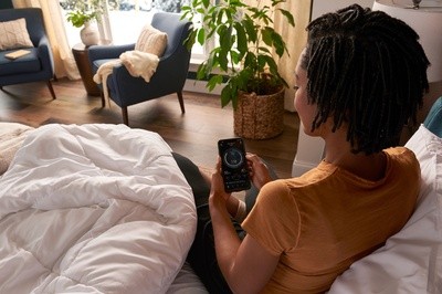 woman sitting on her mattress while looking at her Sleep Report from Tempur-Pedic Sleep Tracker AI with a house plant and chairs in the corner